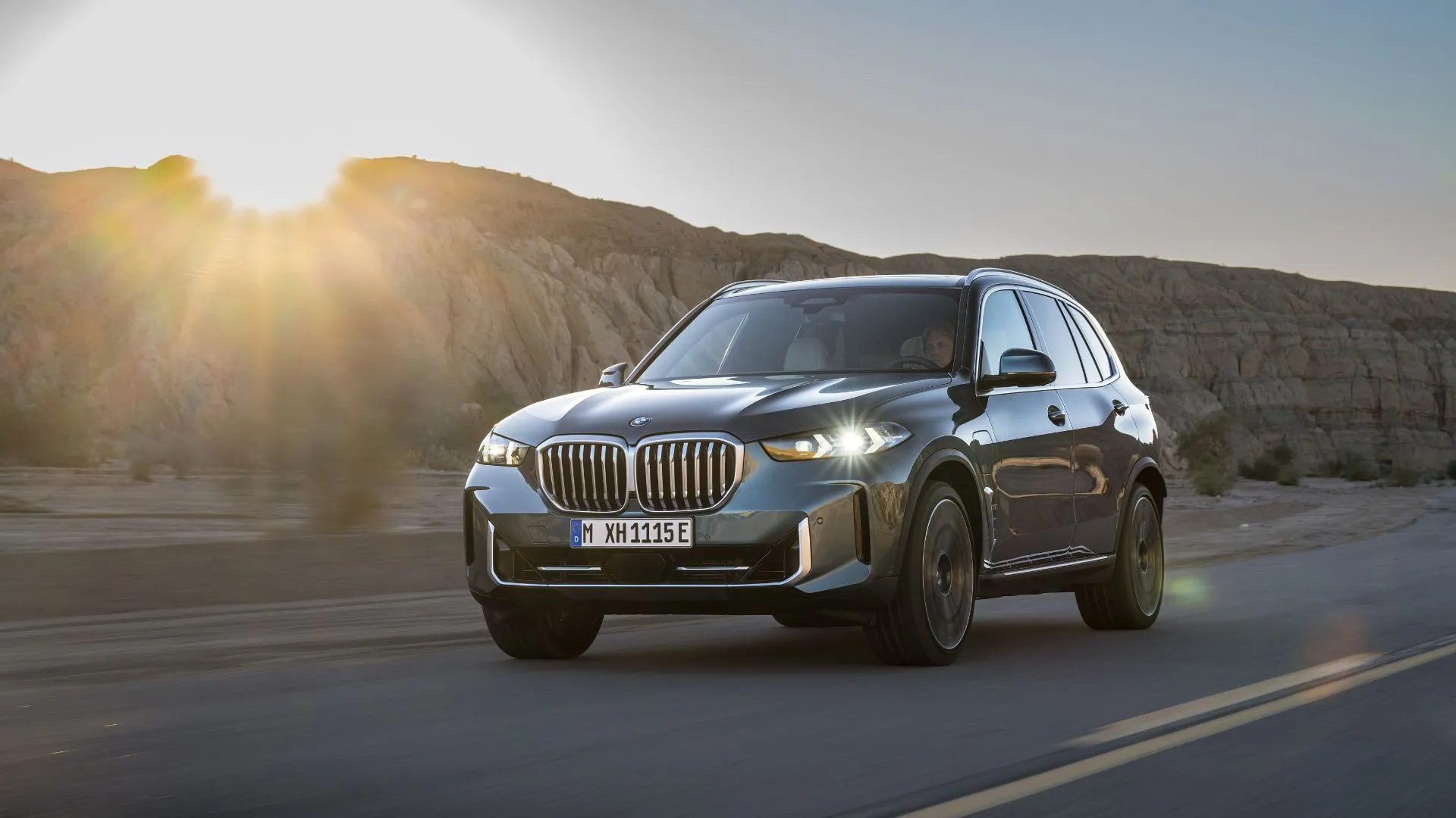 BMW X5 xDrive50e 2024 màu xanh dương đang lái trên đường nông thôn vào lúc hoàng hôn, thể hiện sự mạnh mẽ và sang trọng của dòng SUV