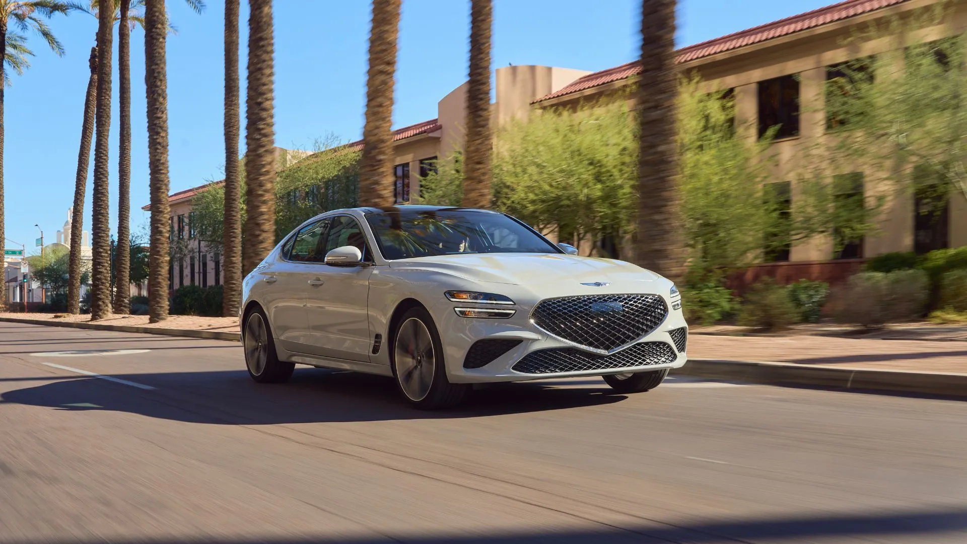 Genesis G70 2025 màu xám đang lướt đi trên đường phố ngập nắng, hai bên hàng cây cọ tạo nên khung cảnh sang trọng