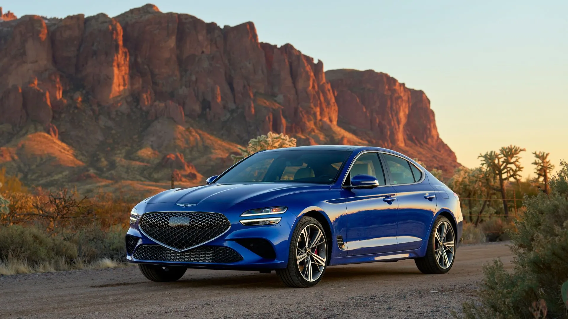Genesis G70 2025 màu xanh dương đỗ trên nền sỏi với núi non phía sau, thể hiện sự kết hợp giữa thiết kế hiện đại và hiệu suất thể thao.