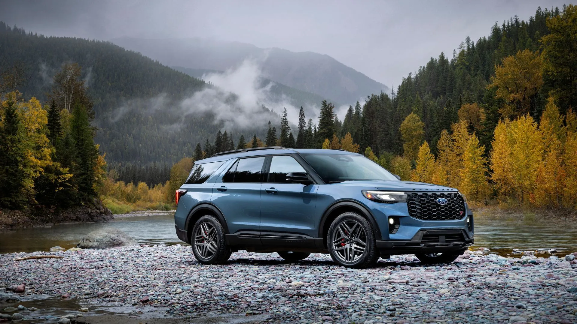 Góc nhìn ba phần tư phía trước của một chiếc Ford Explorer ST 2025 màu xanh dương đỗ trên đá ở vùng núi.