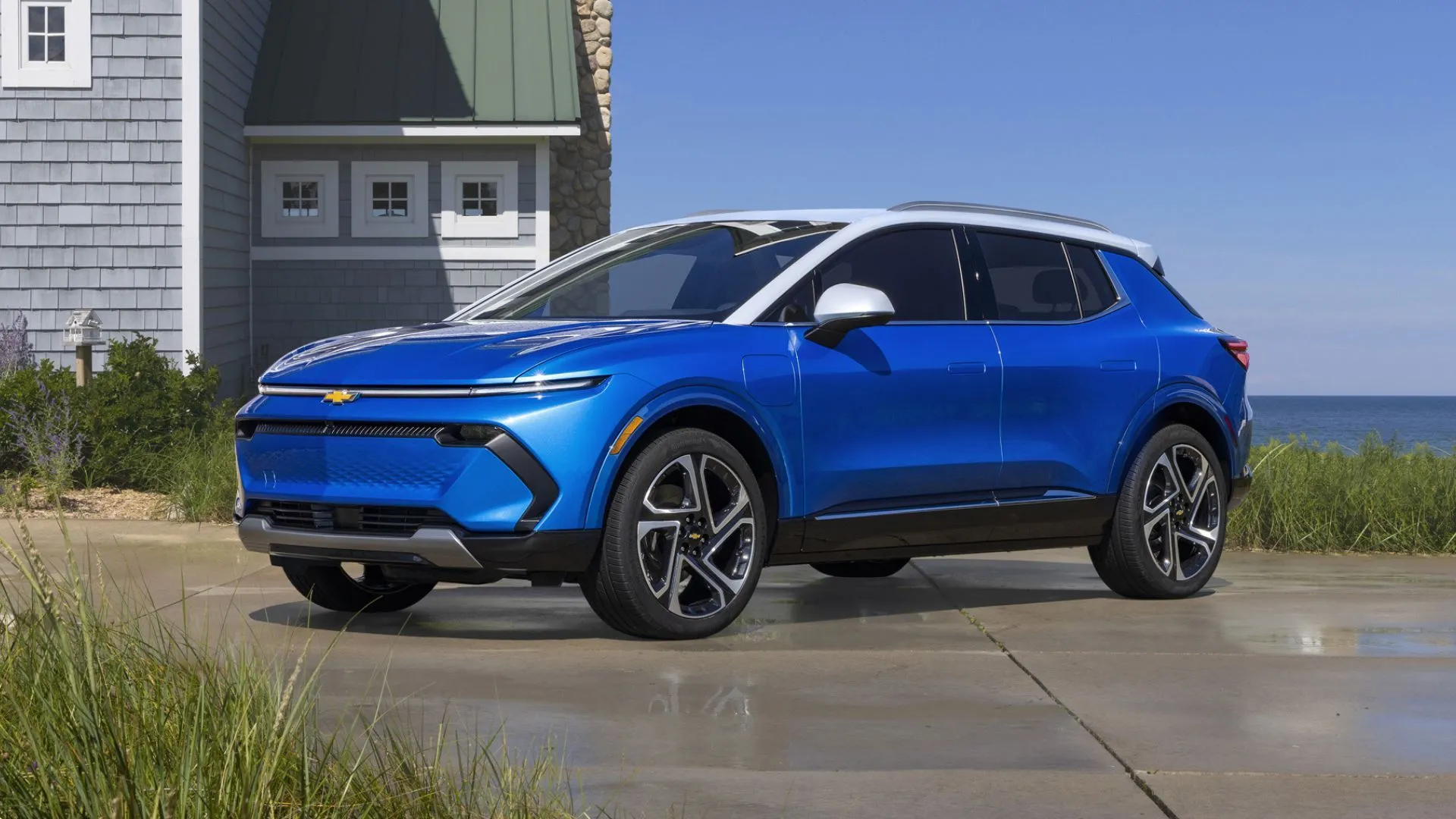 Góc trước của chiếc Chevrolet Equinox EV 2025 màu xanh lam đỗ trước nhà gần bãi biển.
