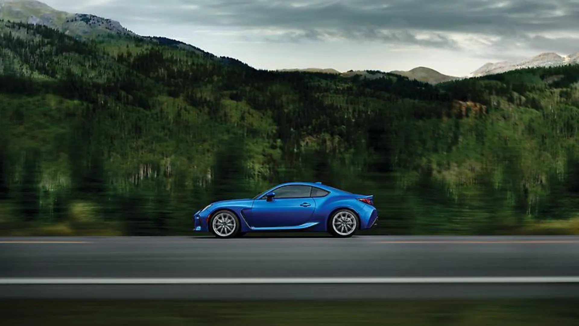 Hình ảnh bên hông của Subaru BRZ 2024 màu xanh đang chạy trên con đường nông thôn với núi và cây xanh phía sau, tượng trưng cho hành trình của mẫu xe này.