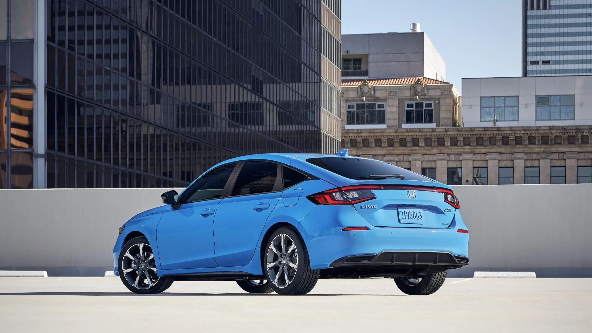 Honda Civic Hatchback 2025 màu xanh lam đỗ trên sân thượng, góc nhìn 3/4 từ phía sau với các tòa nhà cao tầng làm nền.