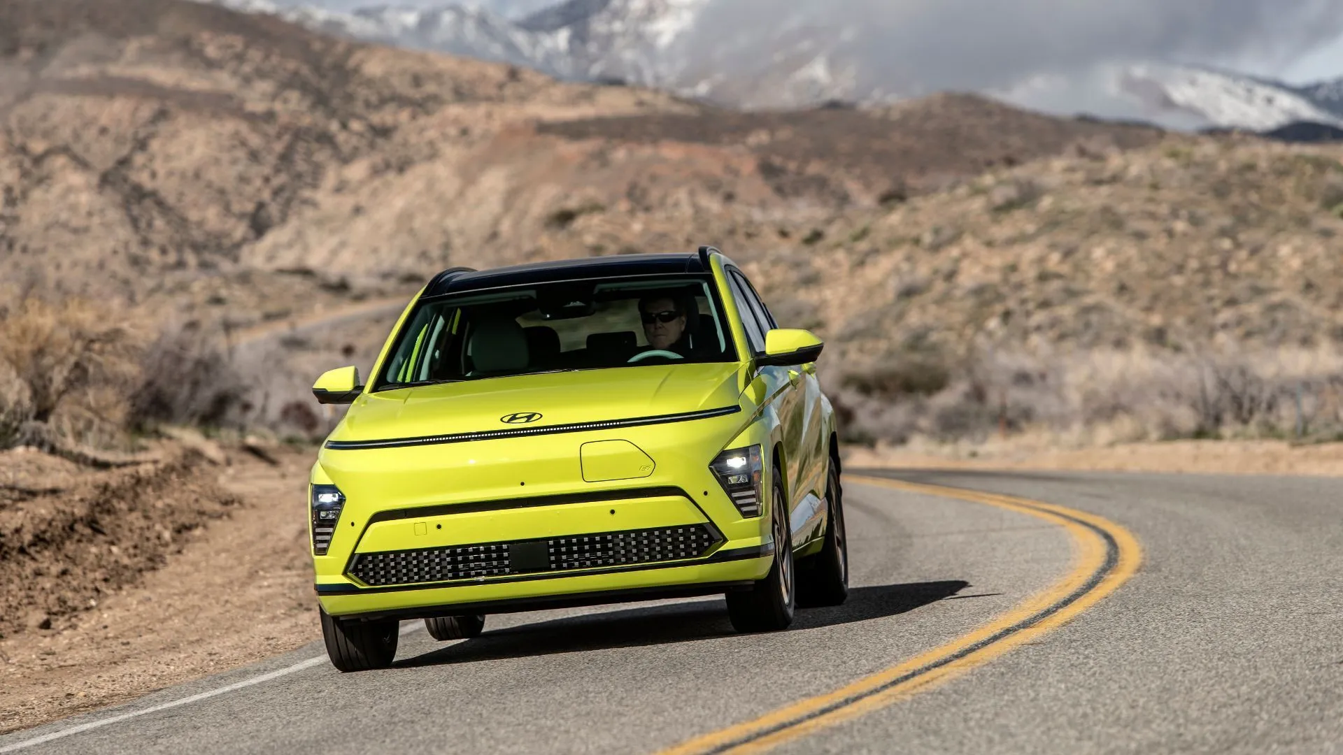 Hyundai Kona Electric 2025 màu vàng đang chạy trên con đường nông thôn uốn lượn, thể hiện sự năng động và phong cách độc đáo của mẫu crossover điện.