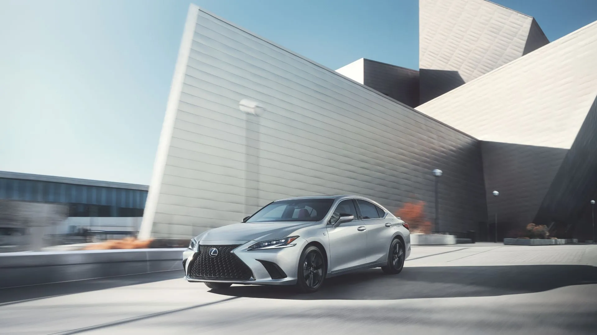 Lexus ES 300h 2023 màu bạc đang chạy trên đường trước một tòa nhà hiện đại, mẫu sedan hybrid sang trọng với khả năng tiết kiệm nhiên liệu ấn tượng.