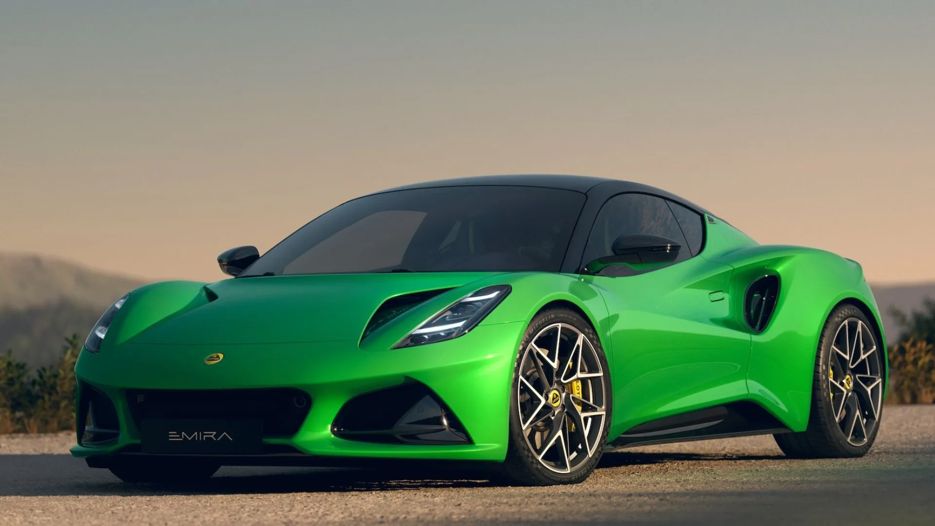 Lotus Emira 2025 màu xanh lá đỗ trên nền sỏi với núi non hùng vĩ phía sau, thể hiện thiết kế xe thể thao