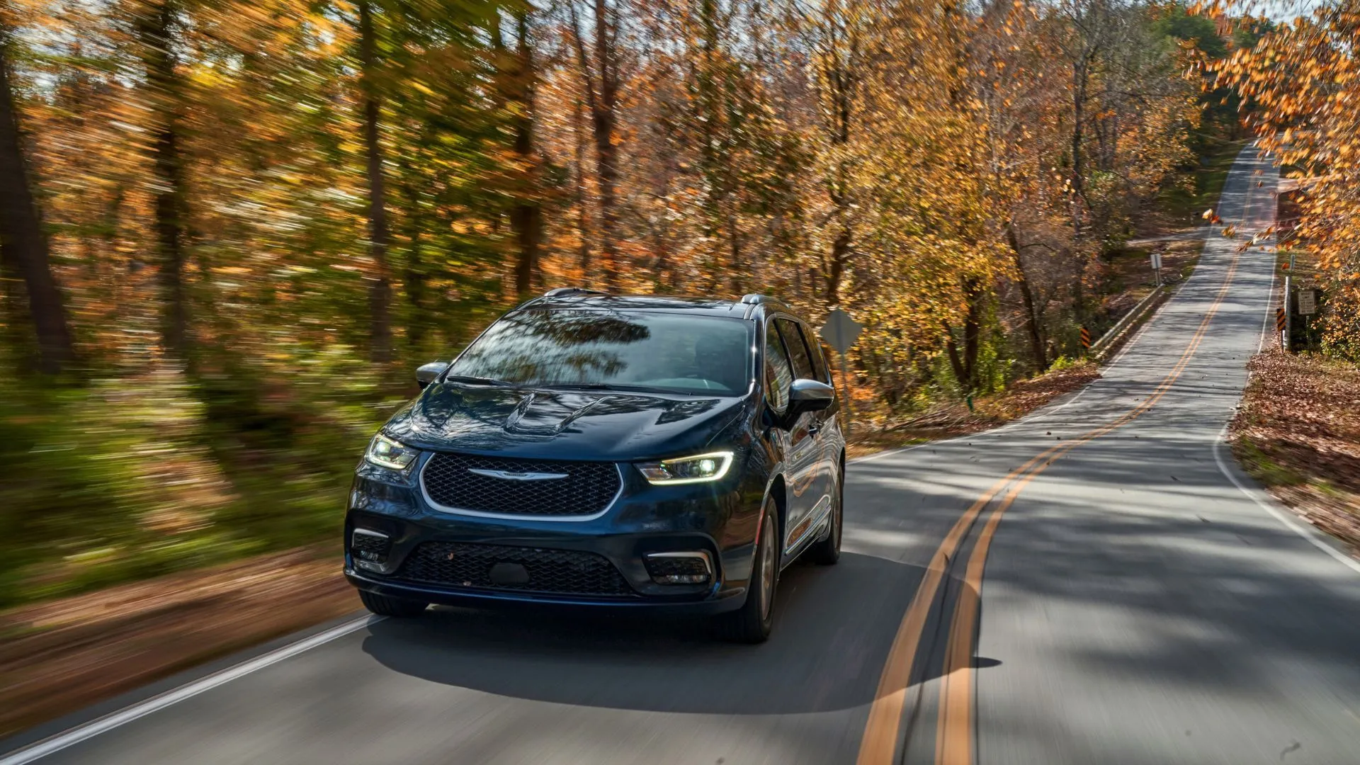 Mẫu Chrysler Pacifica PHEV Pinnacle 2025 màu xanh đậm chạy trên con đường nông thôn rợp bóng cây, nhấn mạnh khả năng di chuyển linh hoạt và vẻ ngoài hiện đại.