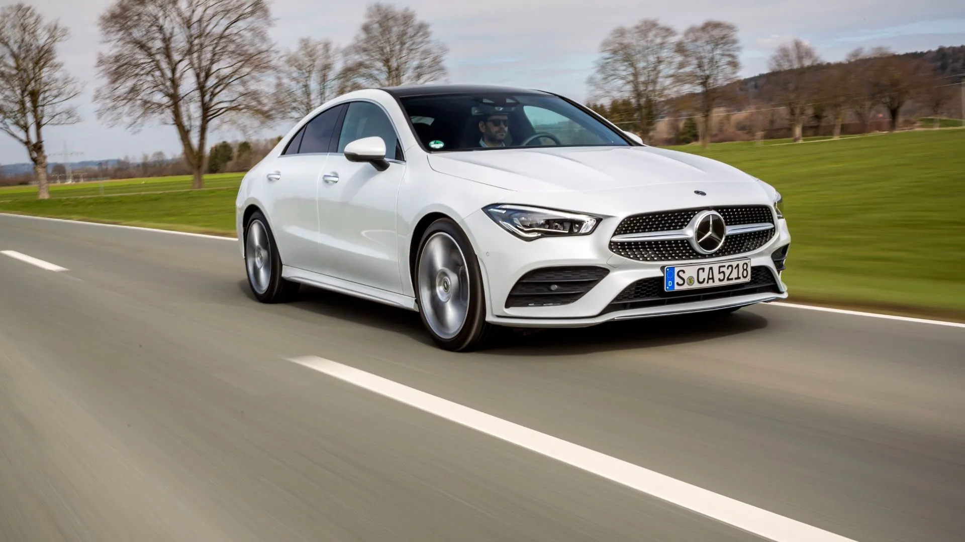Mercedes-Benz CLA 2025 màu trắng di chuyển trên đường quê, thể hiện phong cách coupe và công nghệ MBUX cao cấp.