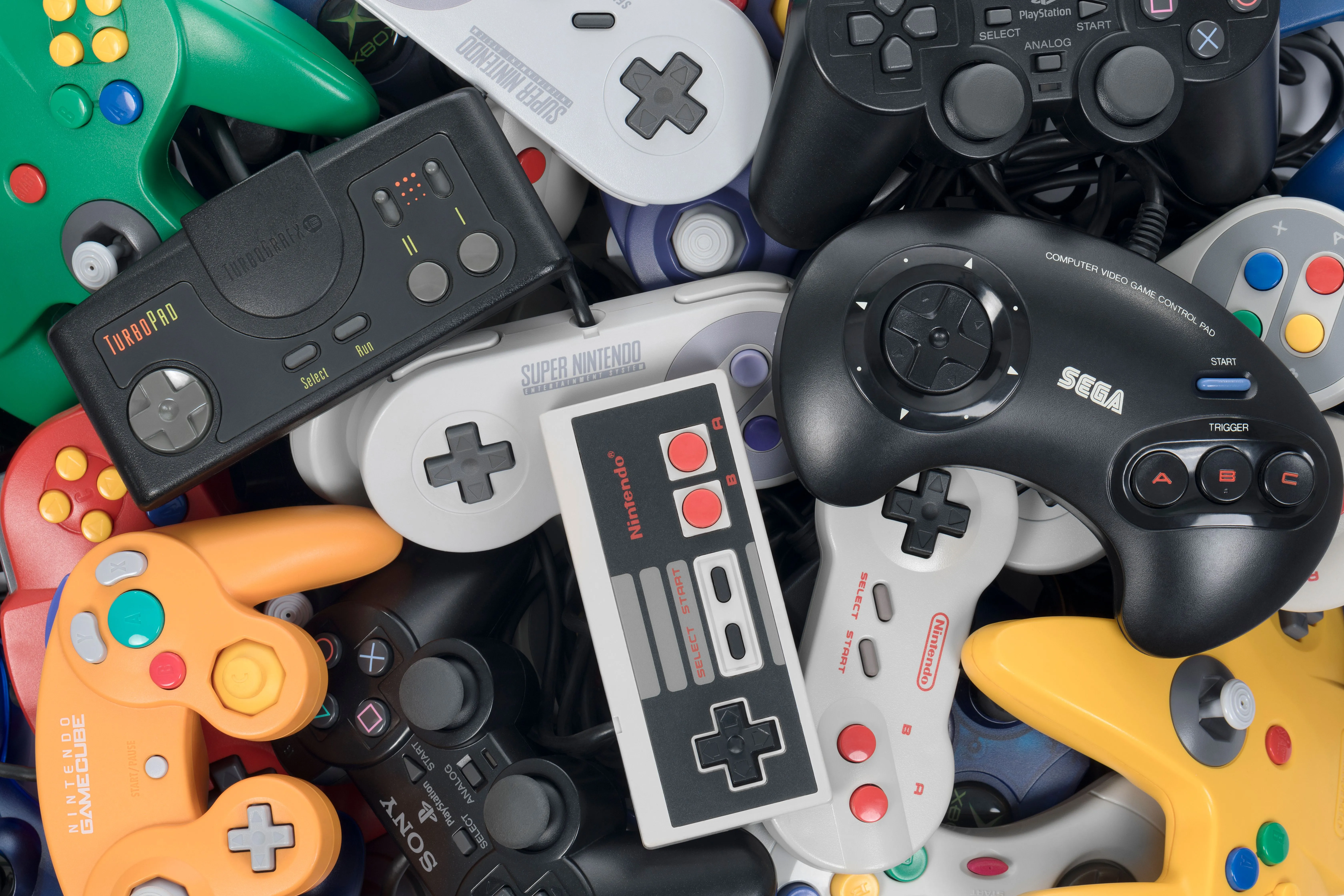 Một chồng các tay cầm chơi game retro từ nhiều thế hệ như NES, SNES và PlayStation, minh họa cho sự phát triển của công nghệ chơi game và nguồn gốc icon tay cầm