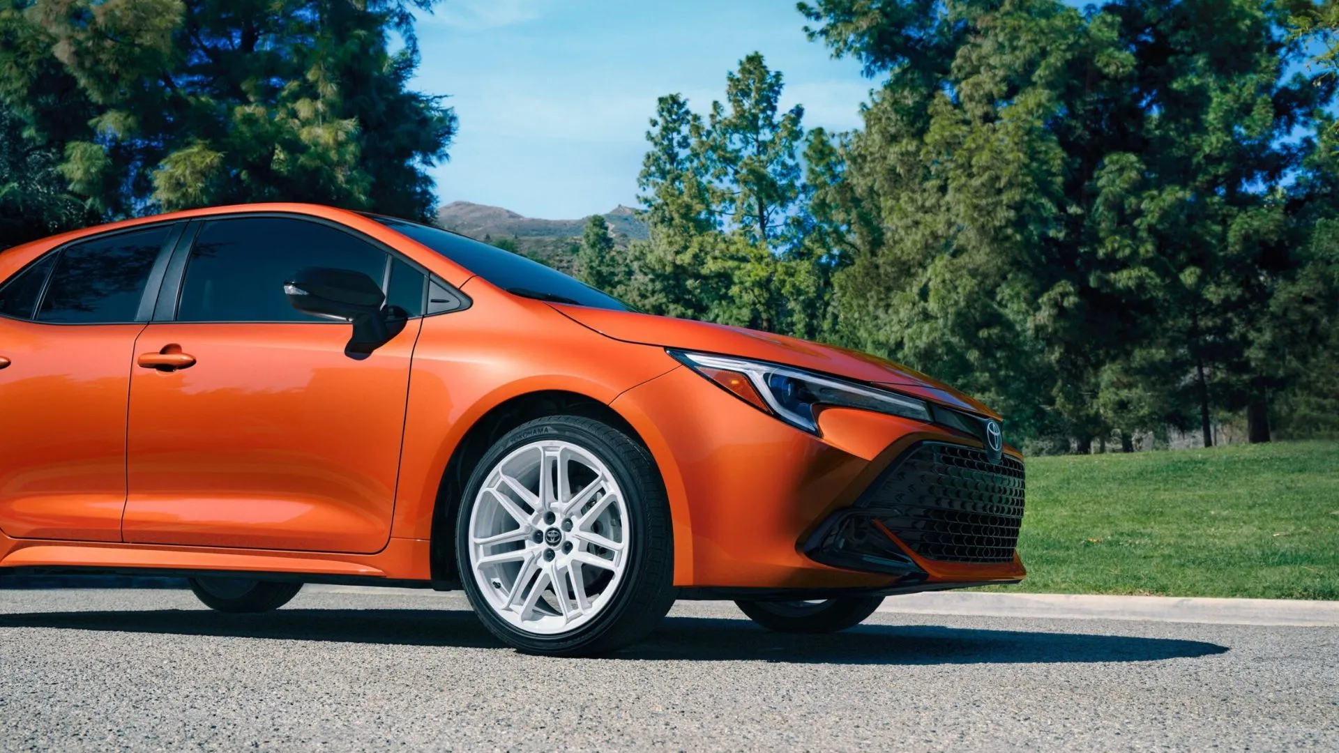 Phần đầu xe Toyota Corolla Hatchback FX Edition 2026 màu cam nổi bật, phía sau là hàng cây xanh.