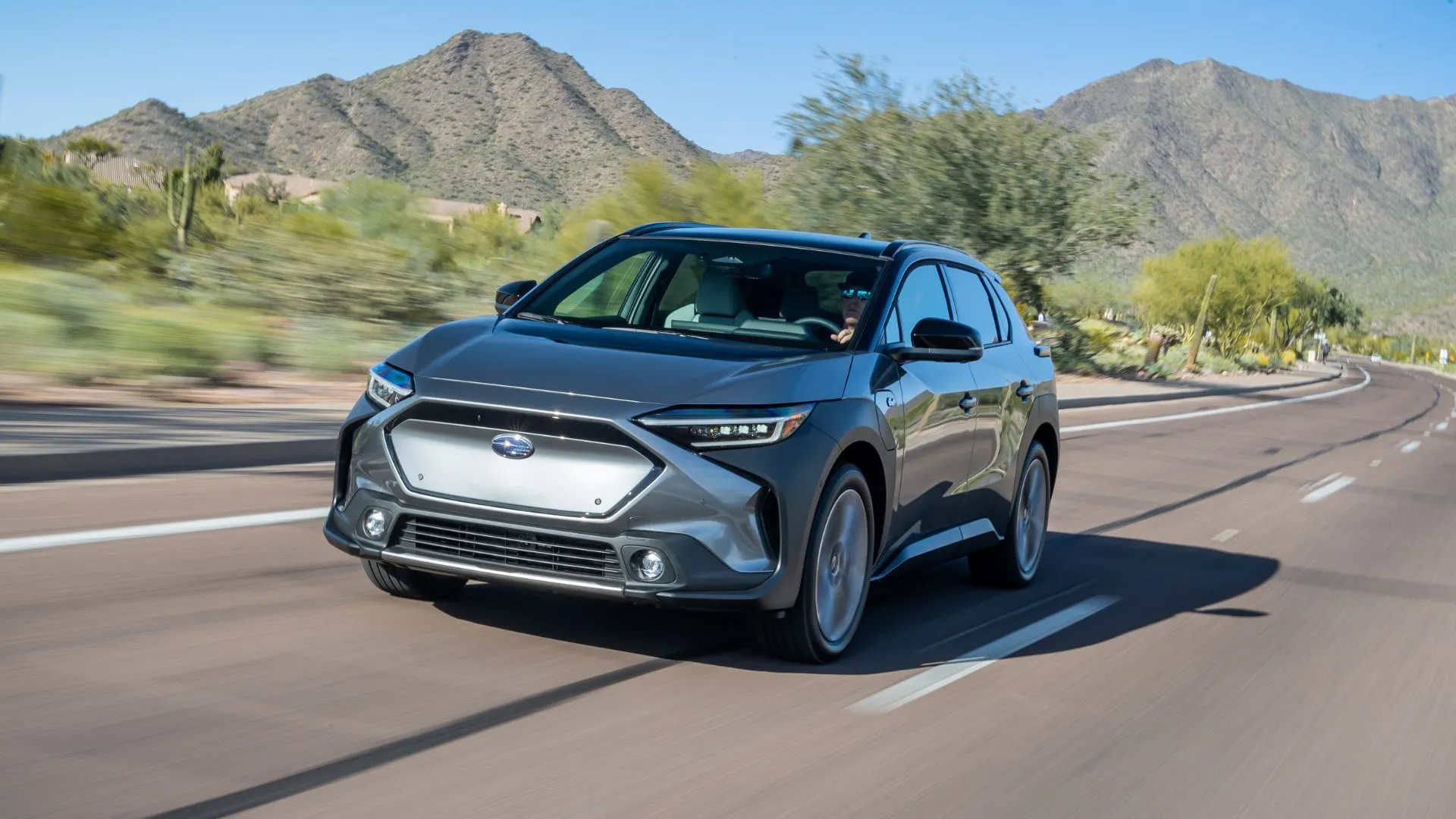 Subaru Solterra 2024 màu xám đang chạy trên con đường nông thôn với núi non phía sau, thể hiện khả năng vượt địa hình và tính bền bỉ.