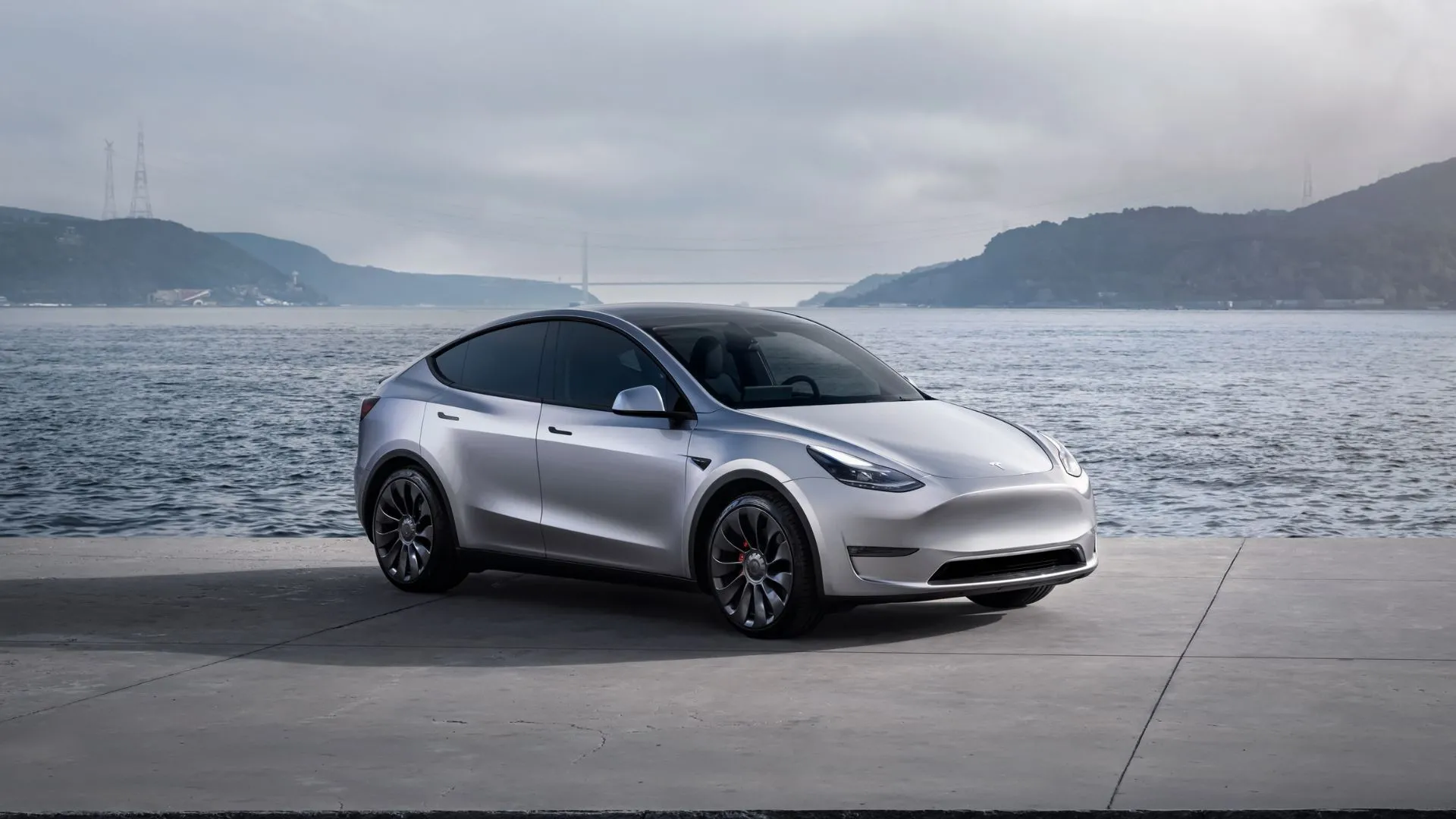 Tesla Model Y màu bạc đỗ bên bờ vịnh với núi non hùng vĩ phía sau, minh họa cho sự bền bỉ và tính thực dụng của dòng xe điện SUV.