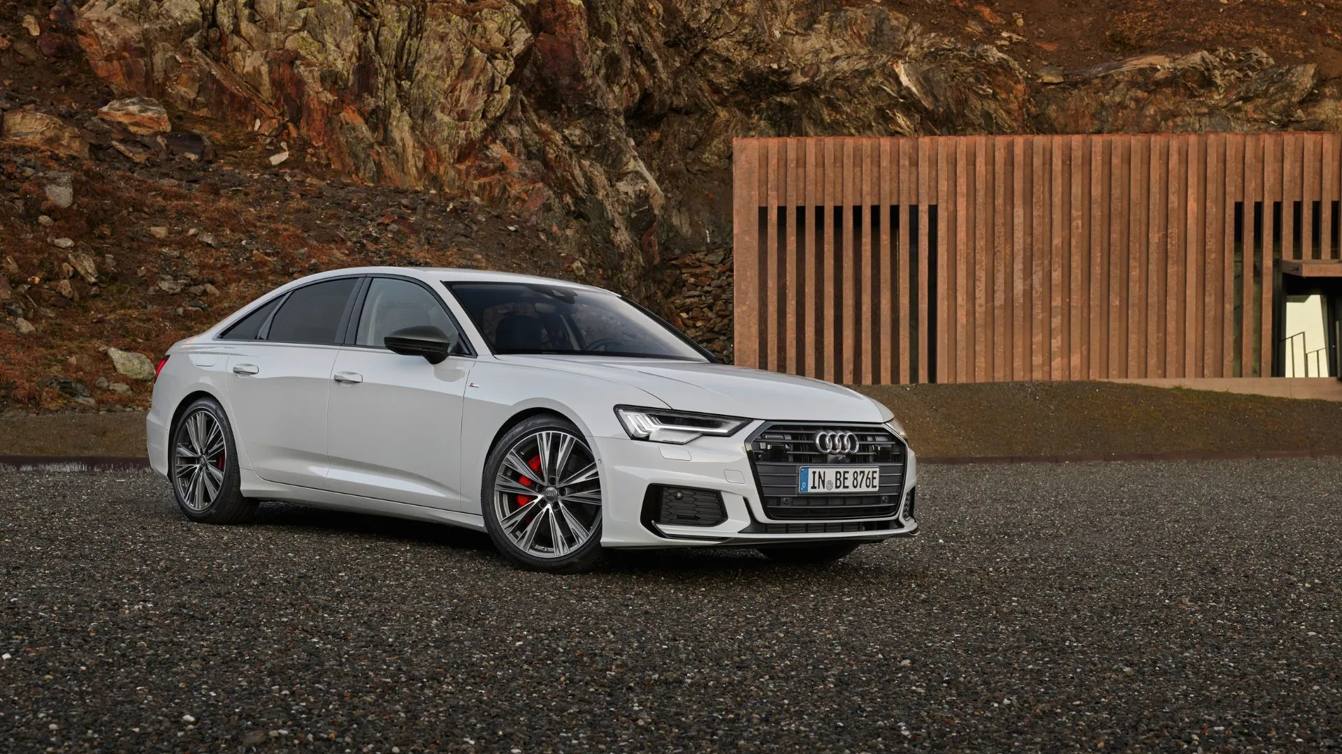 Thiết kế tĩnh, góc 3/4 phía trước của chiếc Audi A6 55 TFSI Sedan màu trắng đỗ trên sỏi với hàng rào gỗ phía sau.