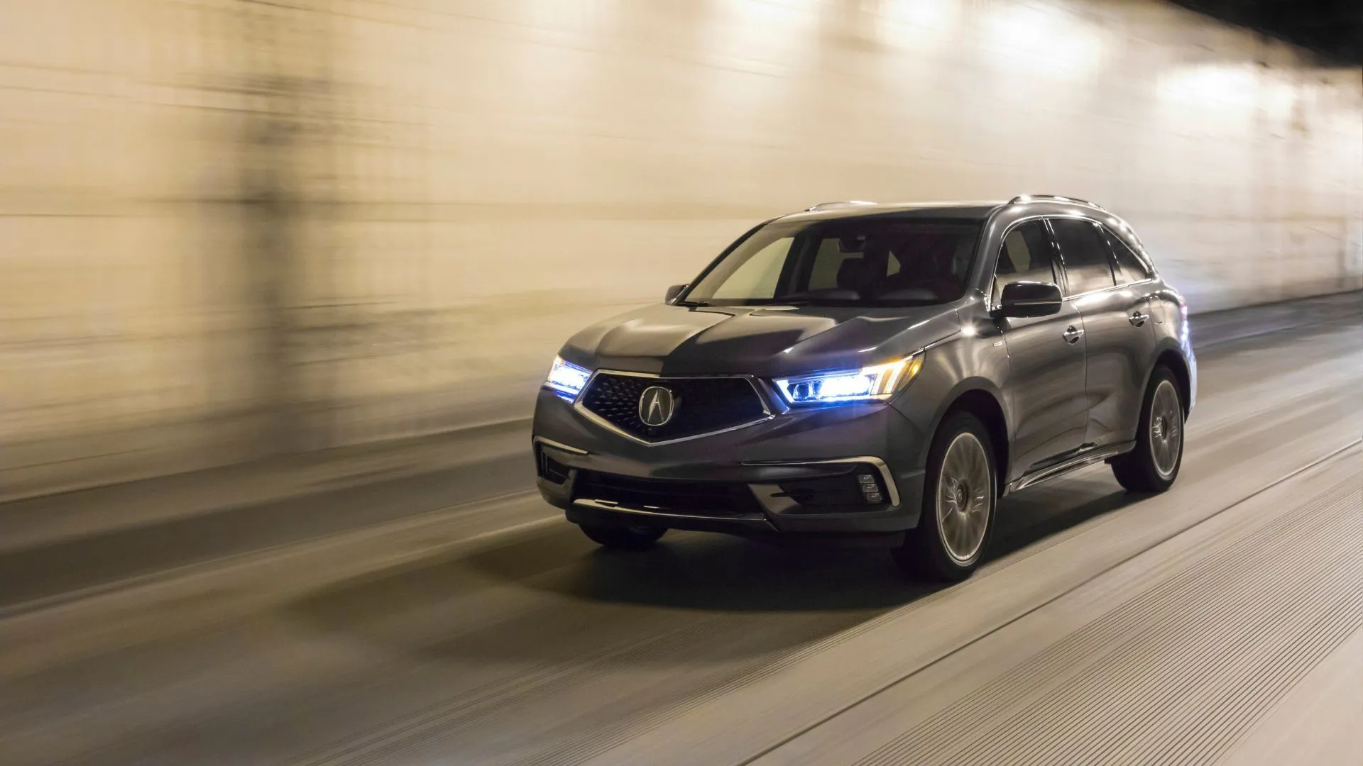Xe Acura MDX Sport Hybrid 2020 màu xám đang di chuyển qua đường hầm với đèn pha sáng, nhấn mạnh tầm quan trọng của hệ thống chiếu sáng an toàn.