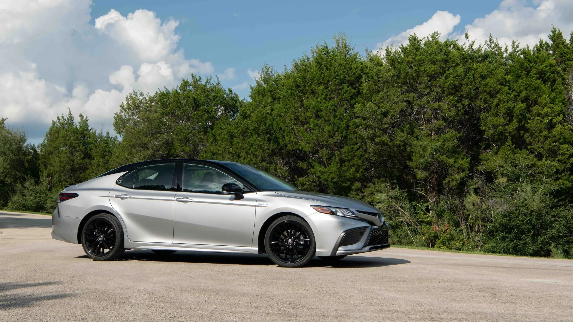 Xe Toyota Camry 2022 Hybrid màu bạc đỗ trên sân nhựa với cây xanh ở hậu cảnh