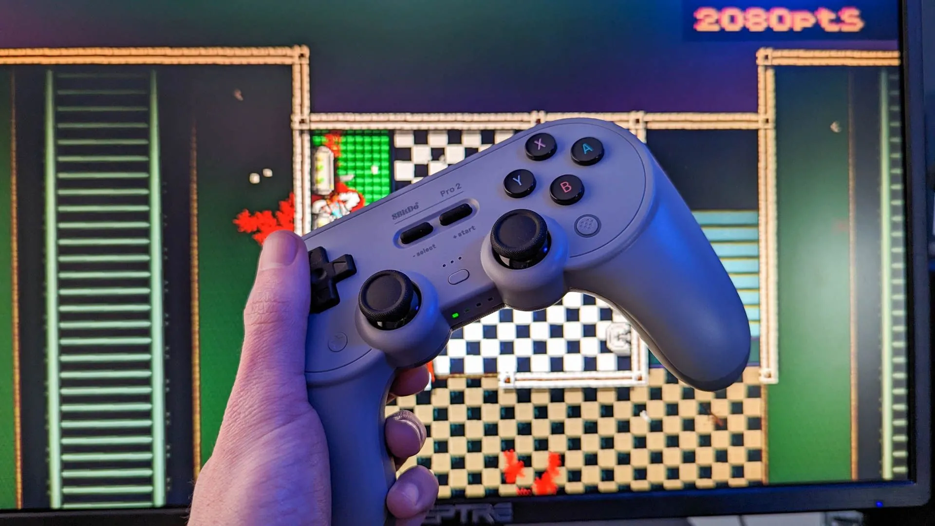 Bộ điều khiển không dây 8Bitdo Pro 2 đặt cạnh màn hình máy tính đang hiển thị game